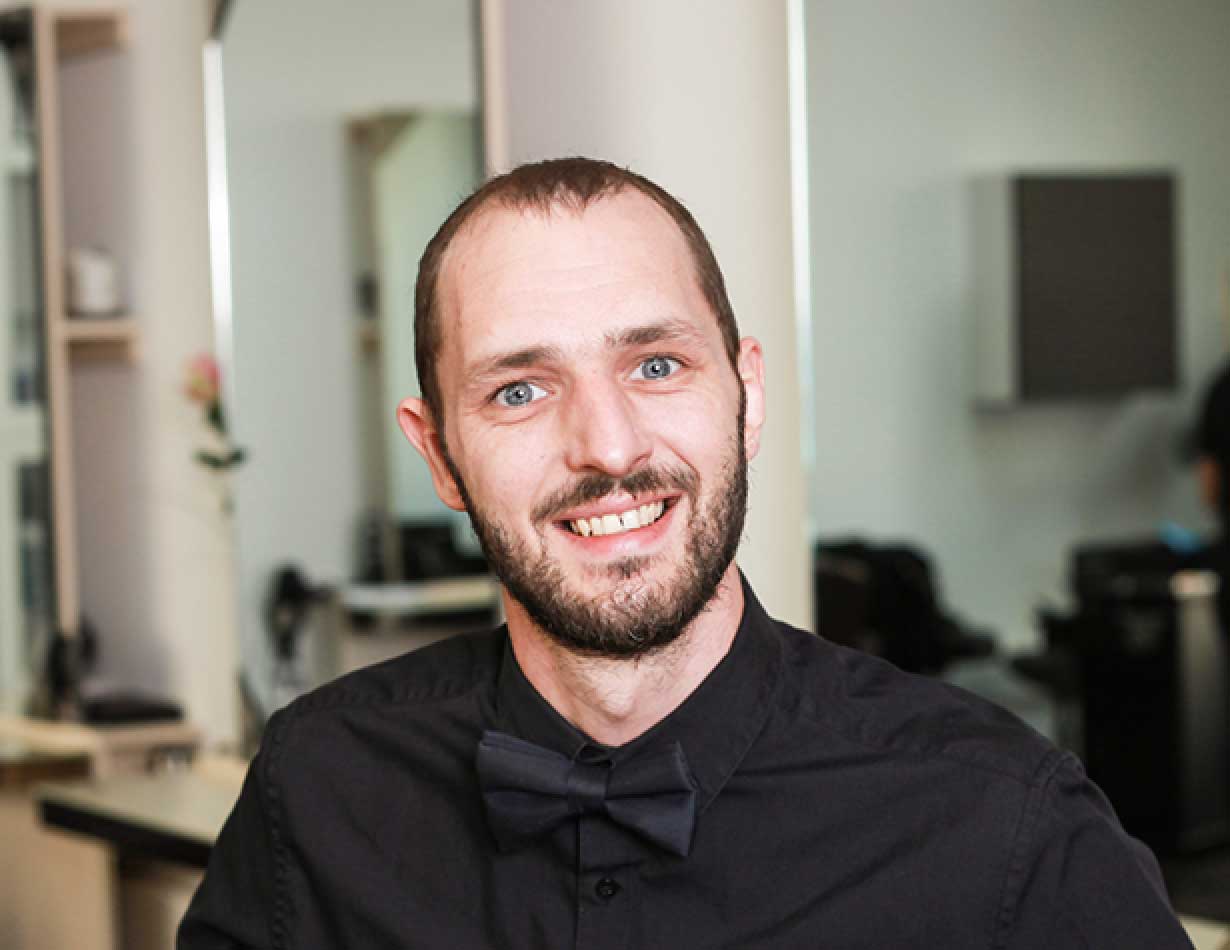 Lächelnder Matthias Lorenz mit kurzem Haar und Bart, gekleidet in schwarzes Hemd mit Fliege, in einem Friseursalon.