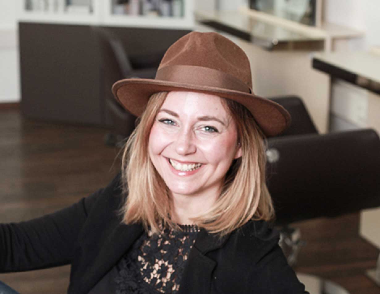 Mandy Mauerer, lächelnd, mit schulterlangen blonden Haaren und braunem Hut in einem Friseursalon.