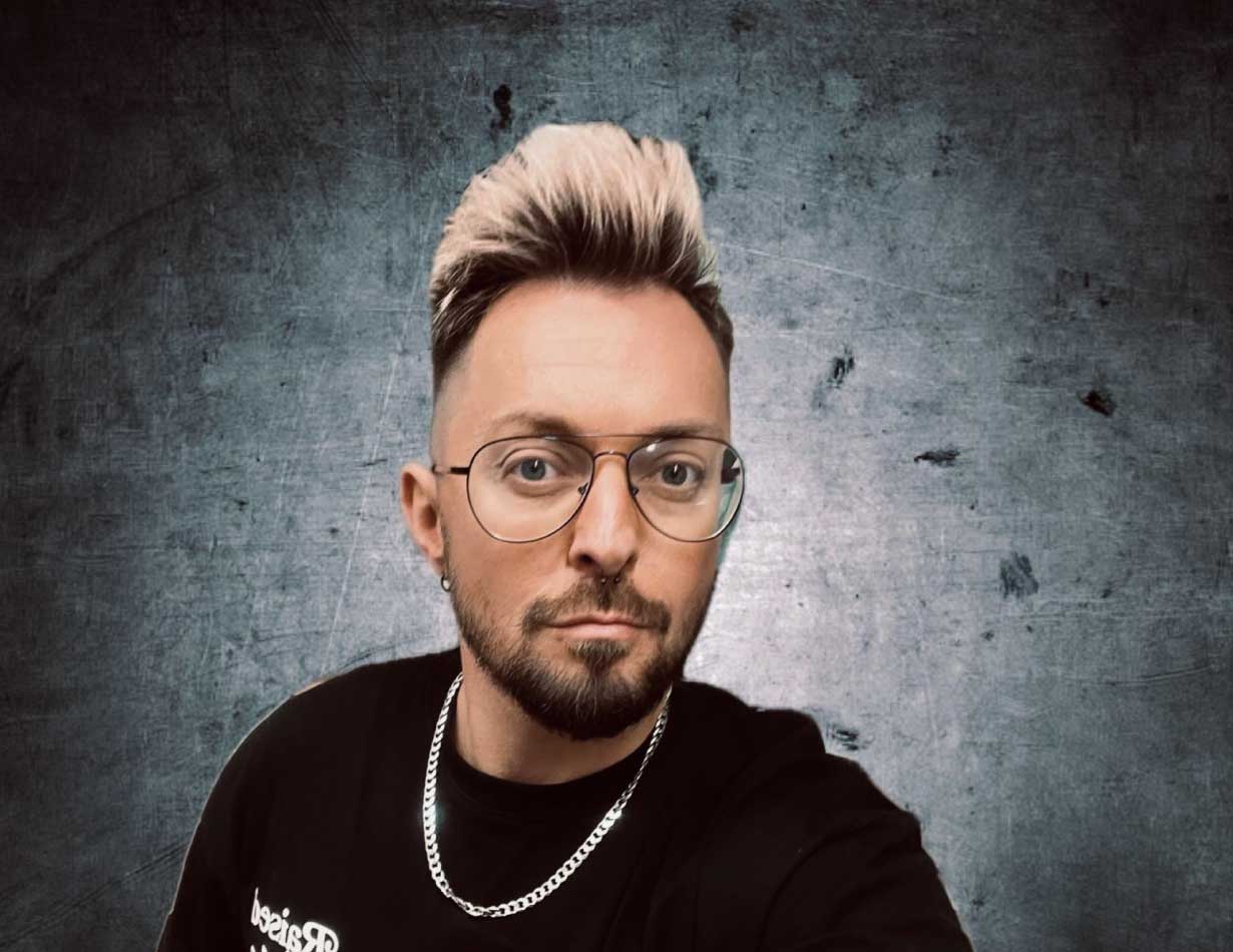 Daniel Meyer mit kurzem, seitlich rasiertem Haar und blond gefärbtem Deckhaar, Bart und Brille vor grauem Hintergrund.